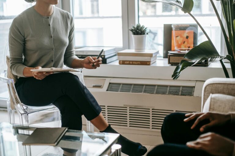 Unrecognizable female psychologist with crossed legs writing information on clipboard during appointment with anonymous ethnic patient in cozy light office